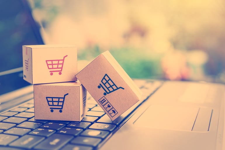 Three little cardboard boxes with shopping cart icon stamped on the side sitting on top of laptop keyboard. Blurred background of a house backyard seen through a window. 