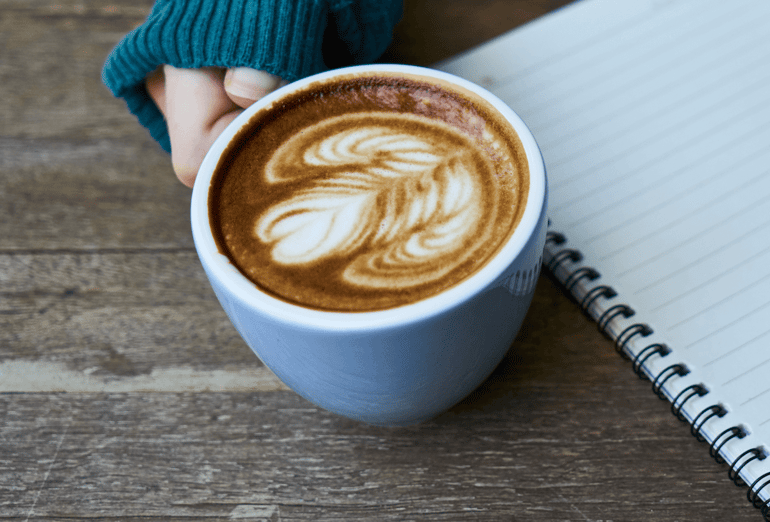 A hand holding a cup of coffee and a notebook on side.