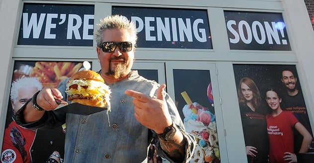 Guy Fieri holding a burger in front of a soon to open restaurant